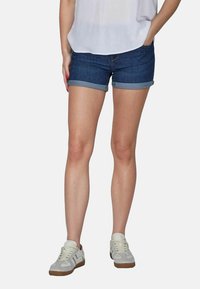 Shorts en denim avec des ourlets retroussés, de couleur bleu foncé, coupe slim, dotés d'une fermeture éclair et d'un bouton à l'avant. Associés à des sneakers blanches décontractées.