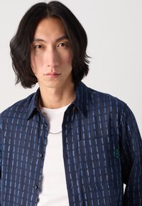 Camisa de color azul marino con un patrón de rayas verticales en azul y blanco, diseño de botones, detalle de bolsillo y un sutil acento de logo verde en el pecho.