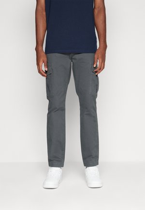 Hombre vistiendo pantalones cargo grises, camisa azul marino y zapatillas blancas, de pie contra un fondo claro y liso.