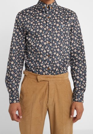 Homme portant une chemise boutonnée à motif floral foncé, rentrée dans un pantalon en velours côtelé marron clair taille haute avec une ceinture en tissu.