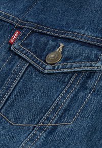 Λεπτομέρεια από τζάκετ τζην που διαθέτει μπλε ύφασμα, μπρούτζινο κουμπί, αντίθετη κίτρινη ραφή και ετικέτα λογότυπου κόκκινου χρώματος στην τσέπη.