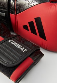 Röda och svarta boxningshandskar med texturerade geometriska mönster och en silverfärgad "COMBAT"-logotyp. Har förstärkt sömnad och skyddande vaddering.
