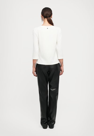 Woman with ponytail wearing a white long-sleeve top and black leather pants, standing facing away against a plain light background.