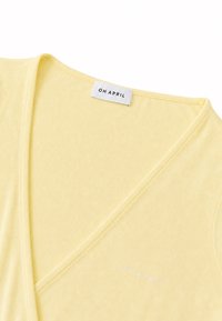 Vêtement jaune clair à col en V avec une étiquette blanche et un petit texte blanc indiquant "OH APRIL" près de l'encolure.