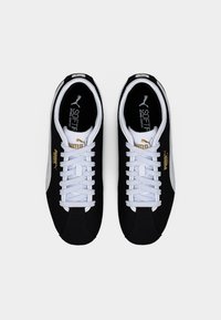 Ζευγάρι μαύρα και άσπρα αθλητικά παπούτσια Puma με λευκά κορδόνια και χρυσές λεπτομέρειες στο λογότυπο, εμφανιζόμενα από πάνω προς τα κάτω σε λευκό φόντο.