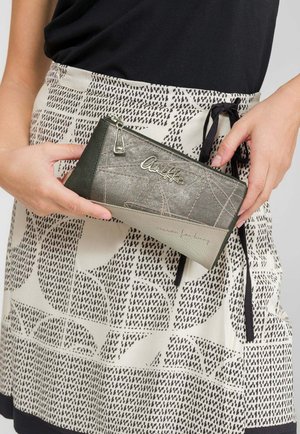 Hände halten eine grau-schwarz gemusterte Reißverschlusstasche mit dem "Anekke"-Logo, getragen über einem beigen Rock mit schwarzem abstraktem Punkt- und Formenmuster.