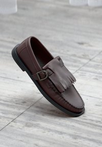 Mocassin en cuir marron avec des détails en franges et une boucle argentée. Présente une semelle en caoutchouc noire contrastante et des bords cousus.