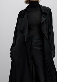 Czarny jedwabny płaszcz trenchowy na dopasowanym czarnym golfie i skórzanych spodniach. Posiada luźną sylwetkę i subtelny połysk.