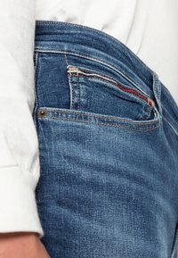 Pantalones vaqueros de mezclilla azul oscuro con un ligero desgaste, costuras de contraste y un pequeño bolsillo delantero. Confeccionados con una tela suave y texturizada.