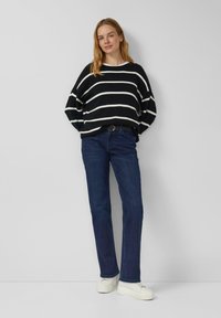 Czarno-biały, szeroki sweter w paski z szerokimi rękawami, zestawiony z ciemnoniebieskimi jeansami o prostych nogawkach i białymi trampkami.