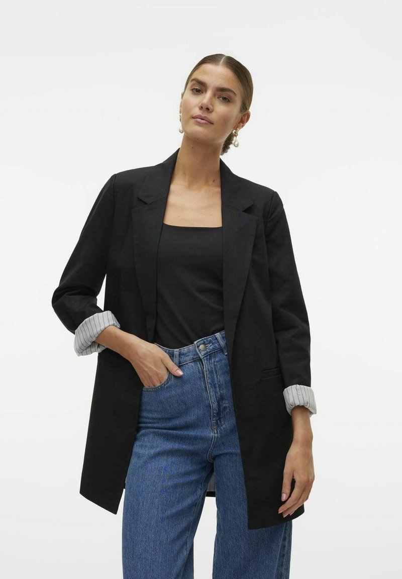 Blazer noir avec un design ajusté, coupe légèrement surdimensionnée, et poignets rayés gris. Porté sur un top noir et associé à un jean bleu.