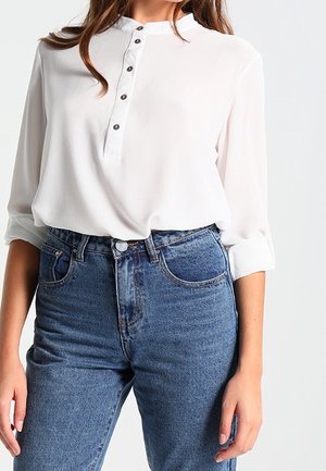 Weißes Hemd mit Stehkragen, ausgestattet mit einer Knopfleiste mit vier dunklen Knöpfen, kombiniert mit hoch taillierten blauen Jeans.