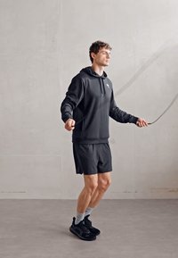 Schwarzer sportlicher Hoodie mit Kängurutasche, kombiniert mit schwarzen Shorts. Die Person trägt schwarze Sneakers und hellgraue Socken und springt Seil.