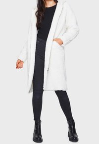 Abrigo blanco con capucha, esponjoso y con una superficie texturizada, abierto al frente y con bolsillos laterales, combinado con jeans negros ajustados y botas negras hasta el tobillo.