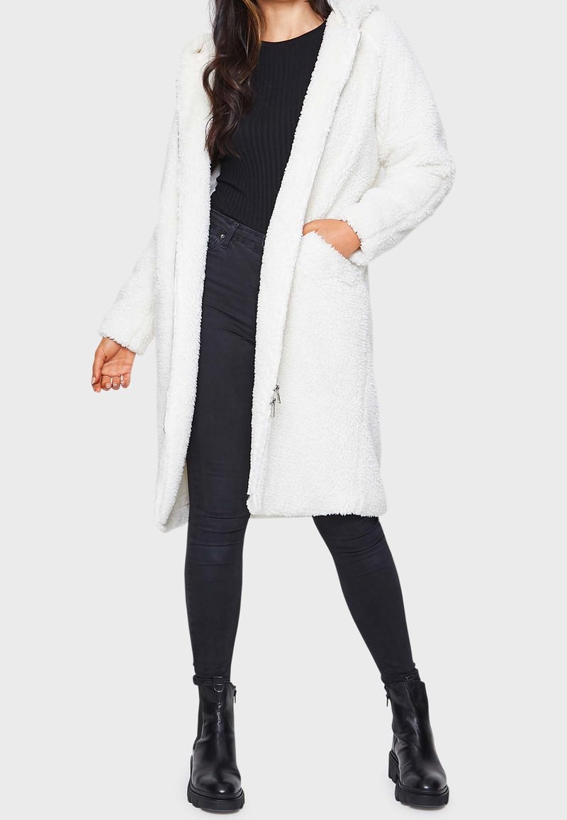 Abrigo blanco con capucha, esponjoso y con una superficie texturizada, abierto al frente y con bolsillos laterales, combinado con jeans negros ajustados y botas negras hasta el tobillo.