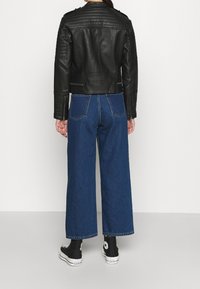 Giubbotto di pelle nera con accenti a costine, abbinato a pantaloni in denim blu a gamba larga. Sneakers alte nere completano il look.