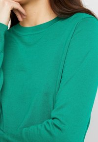 Grön långärmad skjorta i ribbat material, med rund halsringning och åtsittande ärmar. Texturen är slät och något elastisk.
