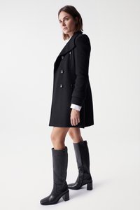Manteau noir croisé en tissu texturé, avec des boutons proéminents et une coupe ajustée. Assorti à des bottes noires en cuir à talons carrés jusqu'aux genoux.
