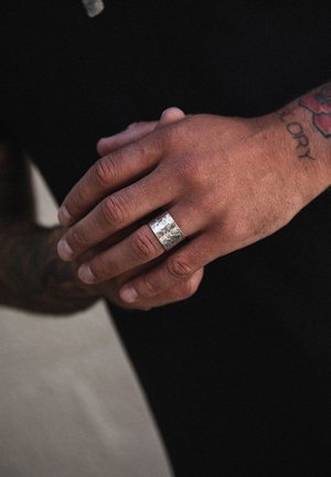Anello d'argento con una finitura martellata, indossato su una mano che mostra tatuaggi. Il tono della pelle contrasta con il metallo liscio e lucido dell'anello.