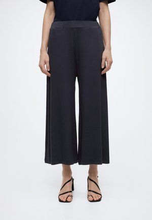 Μοντέλο που φοράει μαύρο παντελόνι φαρδύ και κομμένο στο ύψος του αστραγάλου, συνδυασμένο με μαύρα σανδάλια με λουράκια και τακούνι μπλοκ, στέκεται μπροστά σε λευκό φόντο.