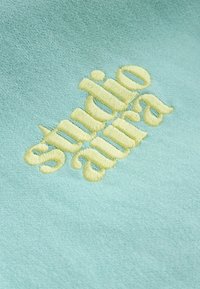 Texte brodé jaune « studio aura » sur fond de tissu bleu clair texturé.