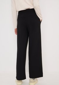 Pantalons larges noirs en tissu doux, avec une taille élastique, deux poches arrière, portés avec des baskets de couleur claire.