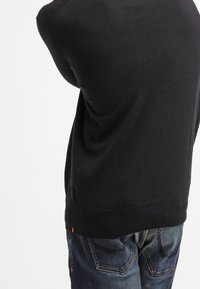 Person, der einen schwarzen Pullover und blaue Jeans trägt, mit erhobenen Armen, von hinten gesehen, vor einem schlichten weißen Hintergrund.