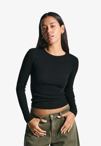 Black long-sleeve top made from soft fabric with a fitted design and round neckline. Paired with olive green trousers, showcasing a casual style.