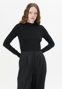 Top negru cu mânecă lungă, cu textură din caneluri și guler înalt. Asortat cu pantaloni negri cu talie înaltă, care au pliseuri frontale și o croială ajustată.