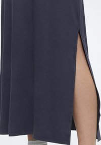 Σκούρο μπλε μίντι φούστα με πλαϊνή σχισμή που αποκαλύπτει το πόδι, συνδυασμένη με ανοιχτογκρι κάλτσες, πάνω σε απλό λευκό φόντο.