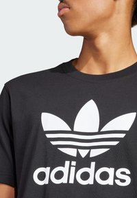 Zwarte katoenen t-shirt met een groot wit Adidas trefoil logo en drie horizontale strepen eronder. Klassiek crew neck ontwerp.