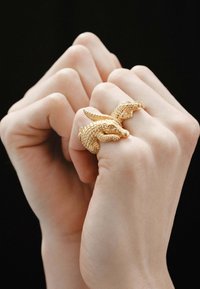 Anillo en forma de serpiente dorada que se enrolla alrededor de dos dedos, con un intrincado detalle en las escamas y una superficie texturada, sobre un fondo oscuro.
