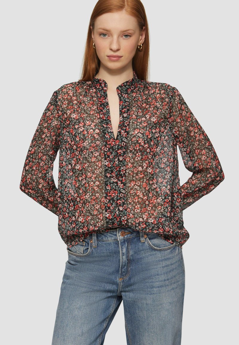 Blouse noire transparente avec un motif floral rouge et blanc, manches longues, coupe décontractée et décolleté en forme de clé. Associée à un jean bleu clair.