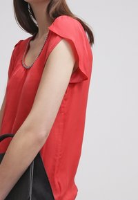 Frau in rotem ärmellosem Blusenshirt mit Perlendekolleté, die eine schwarze strukturierte Handtasche vor einem schlichten hellen Hintergrund hält.