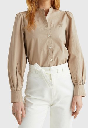 Beige Bluse zum Knöpfen mit puffigen langen Ärmeln und gesticktem Logo, kombiniert mit hoch taillierter weißer Hose und Stoffgürtel, der zu einem Knoten gebunden ist.