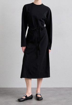 Person trägt ein schwarzes Midikleid mit langen Ärmeln und einem Taillenband sowie schwarze flache Schuhe mit weißen Logobändern und steht auf einem grauen Boden.