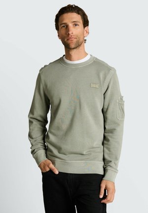 Mężczyzna o krótkich kręconych włosach, ubrany w jasnooliwkową bluzę z kieszenią na rękawie i czarne spodnie, stojący z jedną ręką w kieszeni.