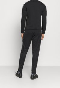 Svart atletisk träningsdräkt med kontrasterande vita sidostråkar; mjukt tyg, figurnära design, ribbade ärmslut och midja, i kombination med svarta sportskor.