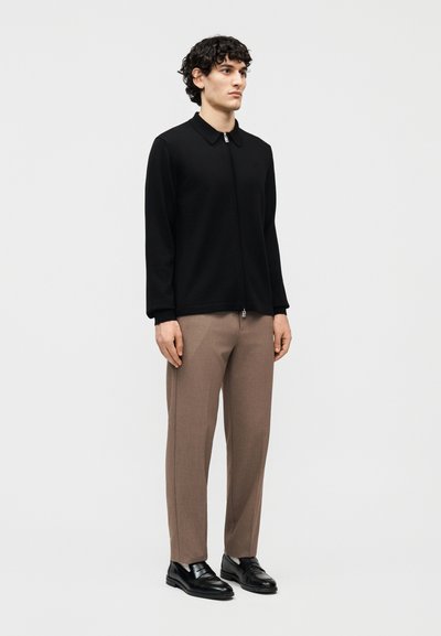 Άνδρας που στέκεται, φοράει μαύρο μπουφάν με φερμουάρ, ανοιχτό καφέ παντελόνι και μαύρα μοκασίνια, με φόντο απλή ανοιχτή επιφάνεια.