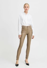 Pantalons skinny en simili cuir beige, taille haute avec passants de ceinture, poches avant et coutures contrastées, associés à une chemise blanche à boutons bien taillée.