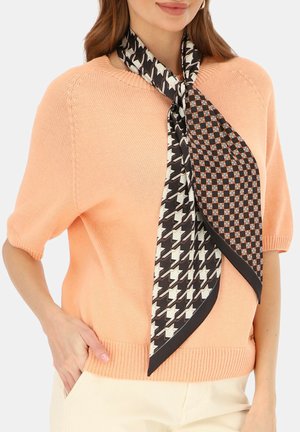 Kobieta ubrana w brzoskwiniowy sweter z krótkim rękawem oraz jedwabną apaszkę w czarno-biały wzór zawiązaną wokół szyi, z ręką w kieszeni.