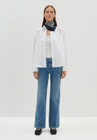 Chemise blanche à manches longues, écharpe rayée bleu marine, débardeur blanc, jeans large bleus clairs et chaussures tressées noires, sur fond uni.