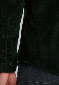 Donkergroen corduroy overhemd met ribbelstructuur, knoopsluitingen en een zijlabel nabij de zoom. Heeft een getailleerde pasvorm en een afgeronde zoom.