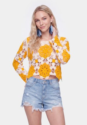 Young woman wearing a white crochet top with large yellow flower patterns and distressed light blue denim shorts, posing with one hand near her face.