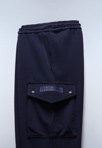 Σκούρο μπλε φόρμα με ελαστική μέση και μια μεγάλη ραμμένη τσέπη στο πλάι με λογότυπο και κούμπωμα με κουμπιά πατιλέτα.