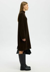 Hnědé oversized svetr šaty vyrobené z texturované tkaniny, s vysokým límcem, dlouhými rukávy a bočními rozparky, doplněné vysokými černými botami.