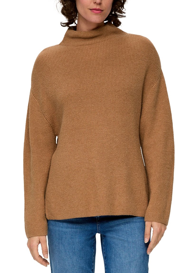 Brauner, grob gestrickter Pullover mit hohem Kragen, lockerer Passform und gerippter Textur, kombiniert mit blauen Jeans. Die Ärmel sind lang und leicht oversized.