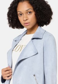 Jasnoniebieska kurtka zamszowa ekologiczna z wyciętym kołnierzem, kieszeniami na suwak i gładką fakturą, zestawiona z białą koszulą z złotymi napisami.