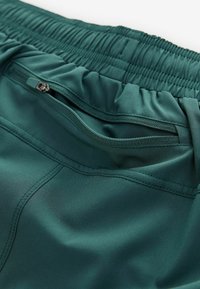 Teal atletiske shorts med elastisk talje. Har en lynlåslomme og glat, let skinnende stof med flade sømme.