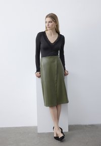Schwarzes Langarmoberteil mit Spitze an der Halspartie, kombiniert mit einem hochtaillierten Rock aus olivgrünem veganem Leder, getragen mit schwarzen hohen Schuhen.